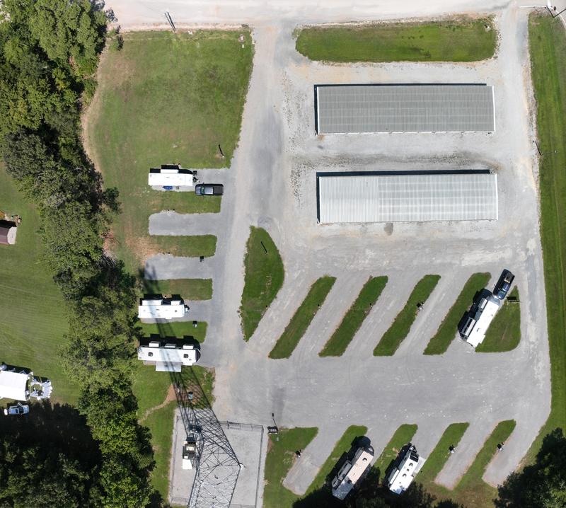 Wilbrook RV Park photographed from directly overhead, north at the top. Wilbrook Street runs along the top of the frame and MS-145, the old highway, down the right-hand side. Two long self-storage buildings stand in the middle of the property. Below them a row of five angled pull-thru pads runs left to right, with a second row of five angled back-in pads below that. A separate column of five straight back-in pads runs down the west side, and a fenced radio tower yard sits at the lower left. Several RVs are parked on the pads, with mown grass strips between every space.