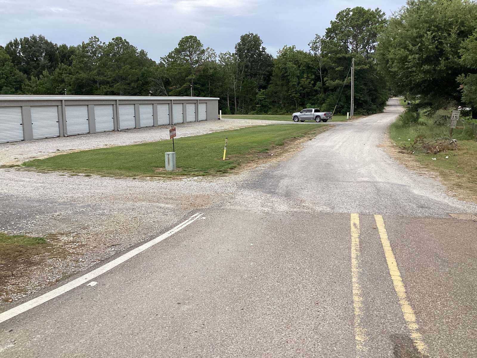 A view down Wilbrook Street toward the park entrance: the long white Colonial Mini Storage buildings run along the left side of the gravel driveway, a silver pickup truck is turning in, and pine trees line the road beyond.