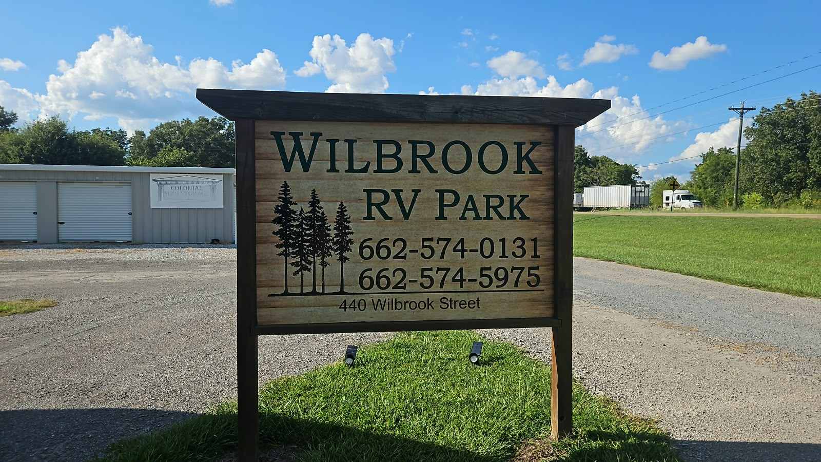 The Wilbrook RV Park sign, seen from the road: a carved wooden board under a timber roof reading WILBROOK RV PARK, with a group of pine trees, the phone numbers 662-574-0131 and 662-574-5975, and 440 Wilbrook Street. The white Colonial Mini Storage buildings stand behind it, and a gravel drive runs past on the right.