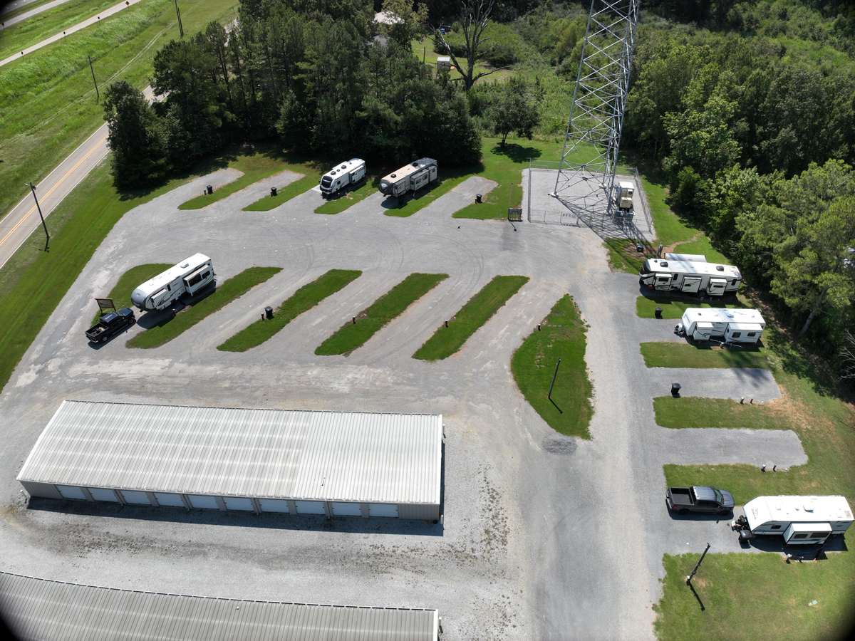 The back-in rows at Wilbrook RV Park from the air, with several travel trailers and fifth wheels parked on the gravel pads. Mown grass strips run between each space, a pickup is parked alongside one RV, and the self-storage buildings and tree line stand behind.