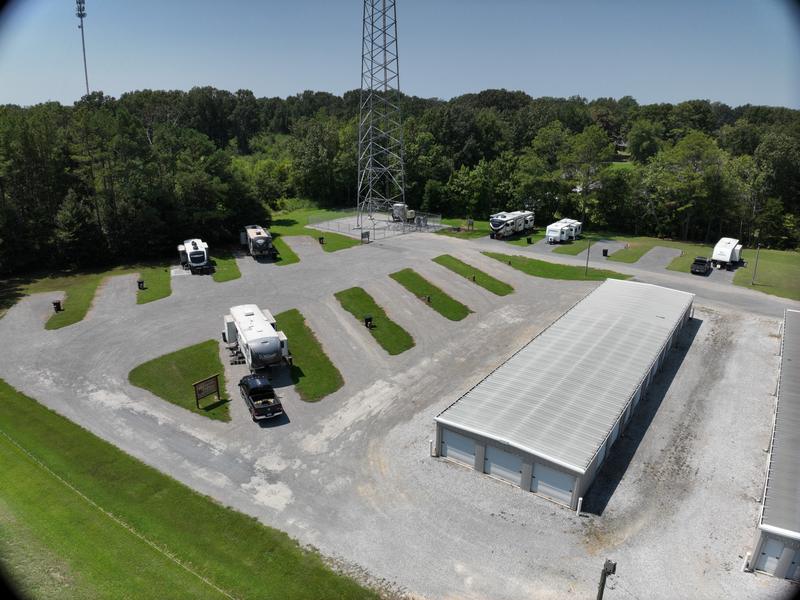 Wilbrook RV Park from the air on a clear summer day, looking across the gravel apron toward the radio tower. The angled pads fan off the apron with mown grass strips between them, several RVs and fifth wheels are parked, and the two long self-storage buildings run along the right-hand side with a tree line beyond.