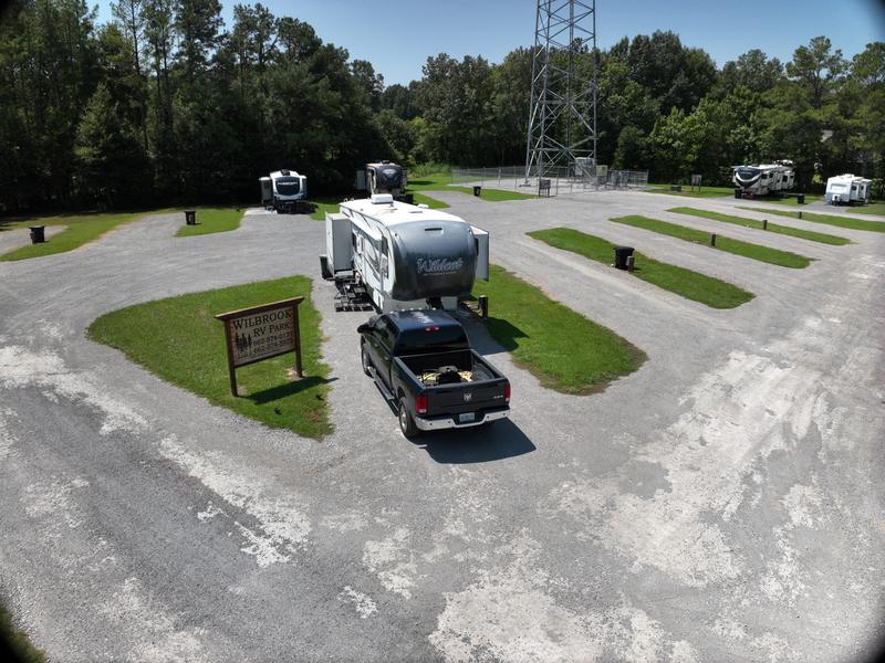 A low aerial view at Wilbrook RV Park with the carved wooden park sign in the foreground, a Forest River Wildcat fifth wheel and a pickup parked together on one pad, empty gravel pads with mown grass strips either side, and the radio tower and tree line behind.