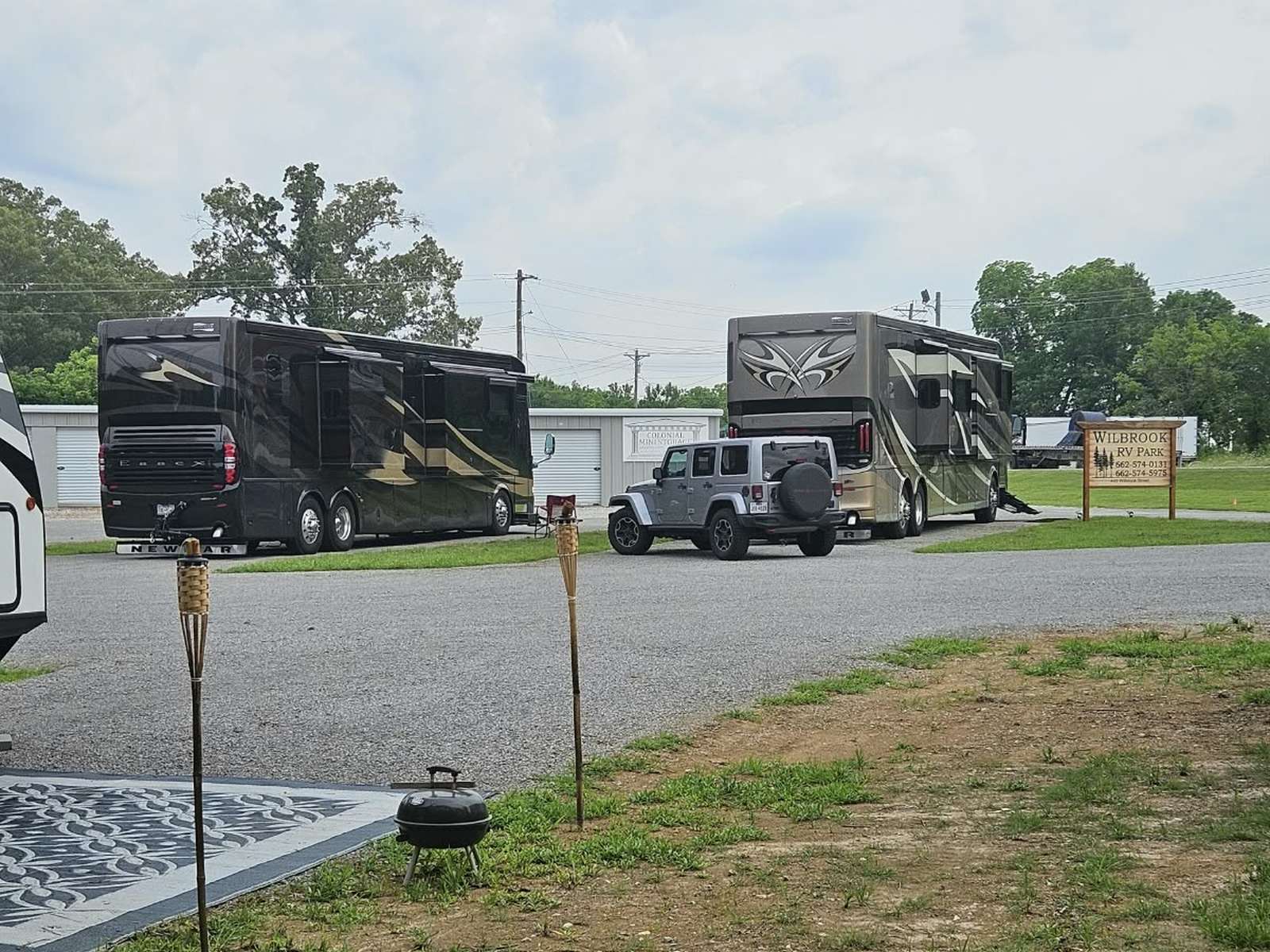 The same two Newmar Essex motorhomes parked in the pull-thru spaces with a Jeep Wrangler towed vehicle between them, the Wilbrook RV Park sign and Colonial Mini Storage building visible behind, tiki torches and a small grill set up at a neighboring site.