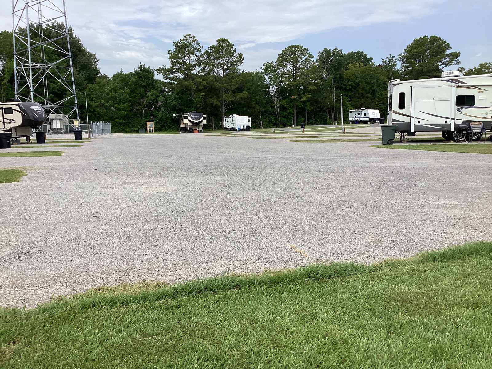 A wide view across the gravel apron at Wilbrook RV Park with several RVs parked on both sides, the cell tower at left and a tree line behind.