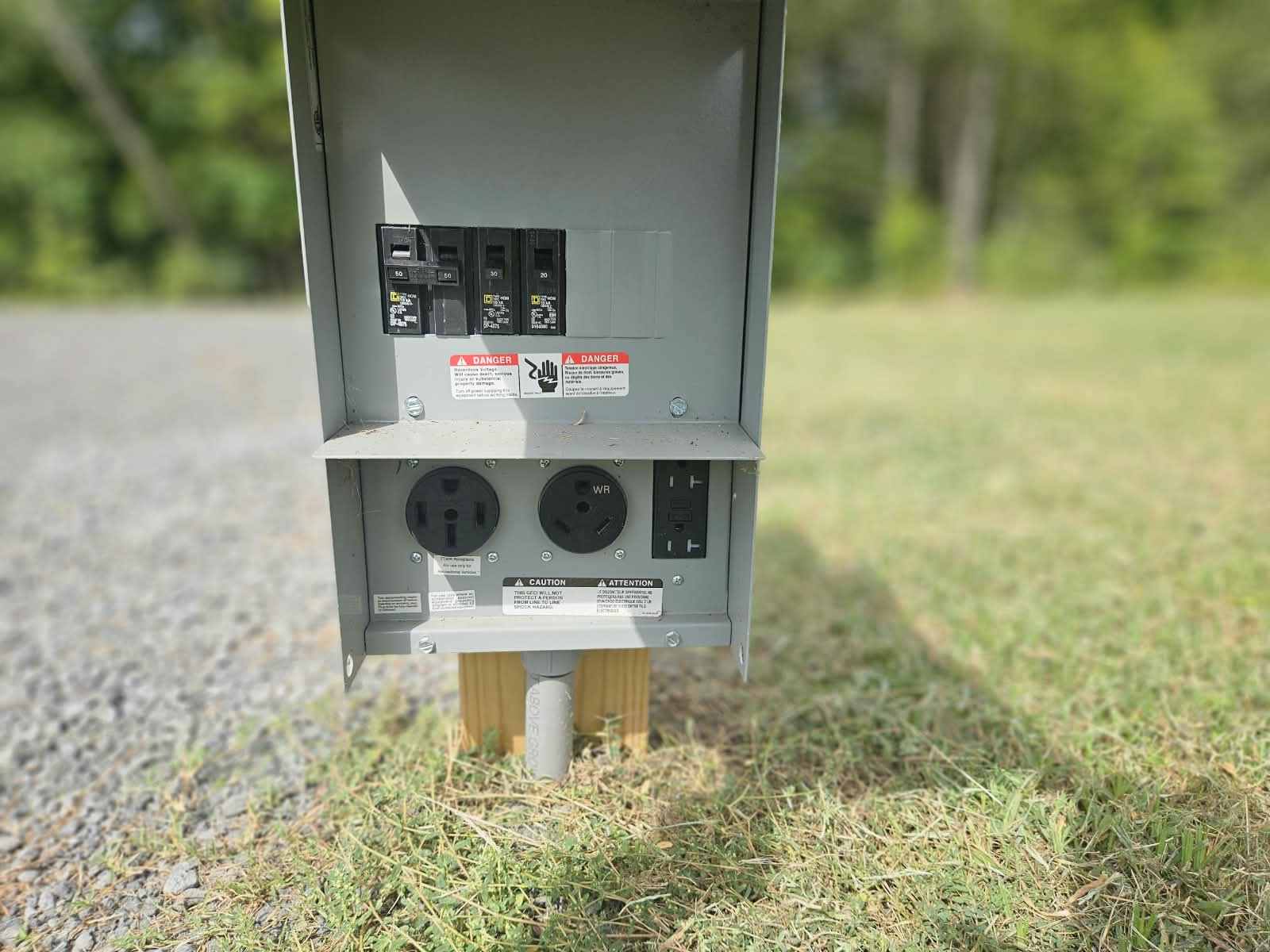 A power pedestal with the panel door open, showing four breakers and, below them, a 50 amp outlet, a 30 amp outlet and a 20 amp GFCI duplex outlet, mounted on a post at the edge of the gravel with grass behind.