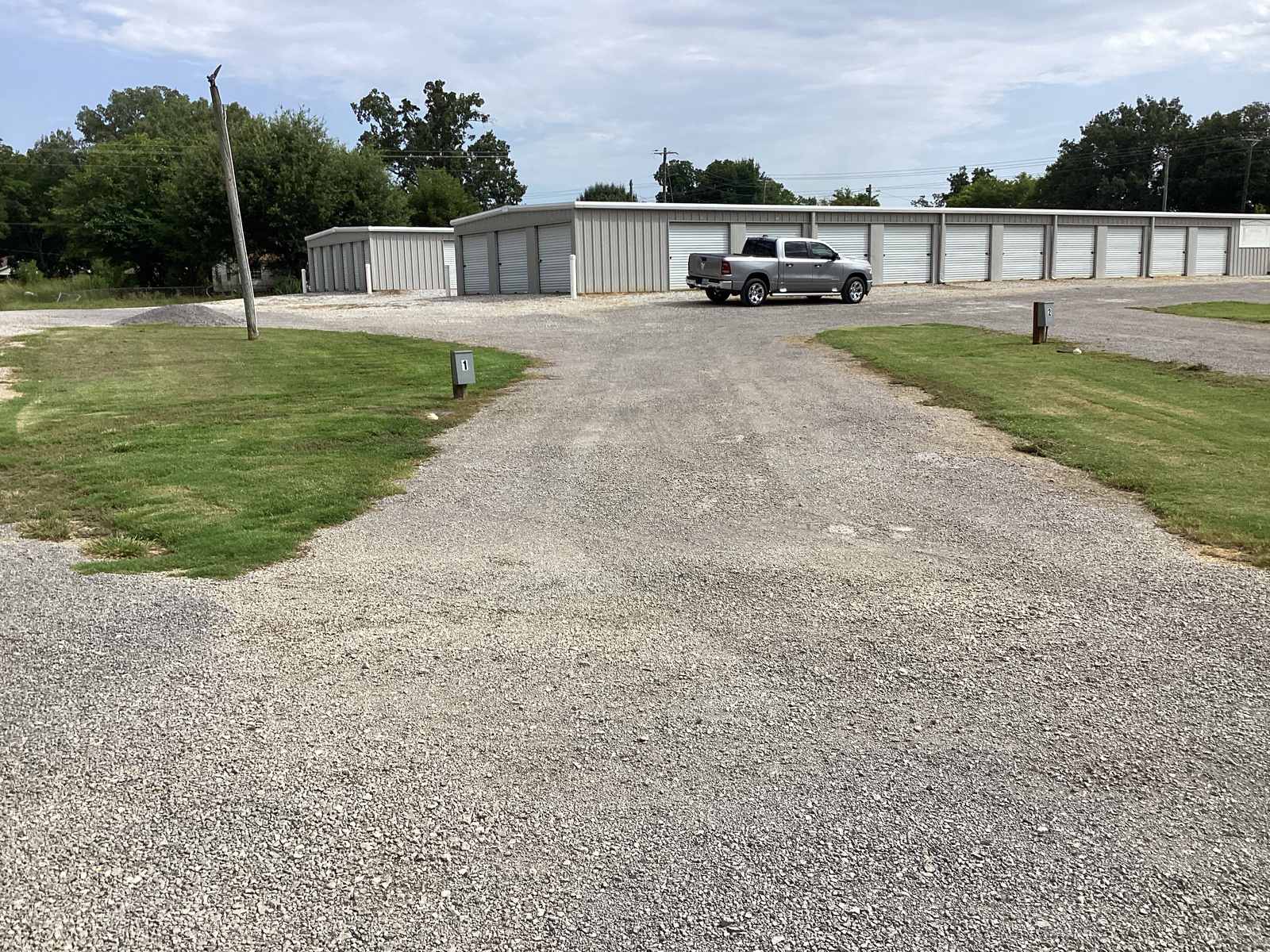 A pull-thru pad photographed down its length, gravel with a grass median splitting the entrance and exit lanes, running past the row of storage buildings.