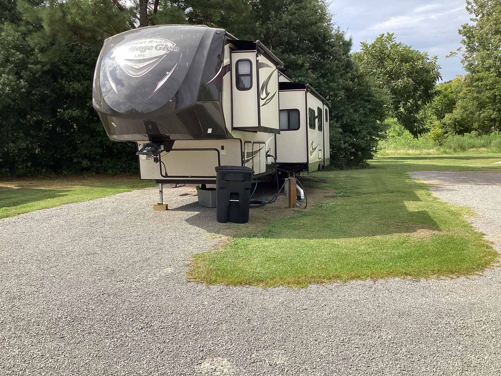 A Forest River Heritage Glen fifth wheel with its awning out, parked on the gravel pad at one of the numbered spaces, mown grass between spaces and mature pine trees behind.
